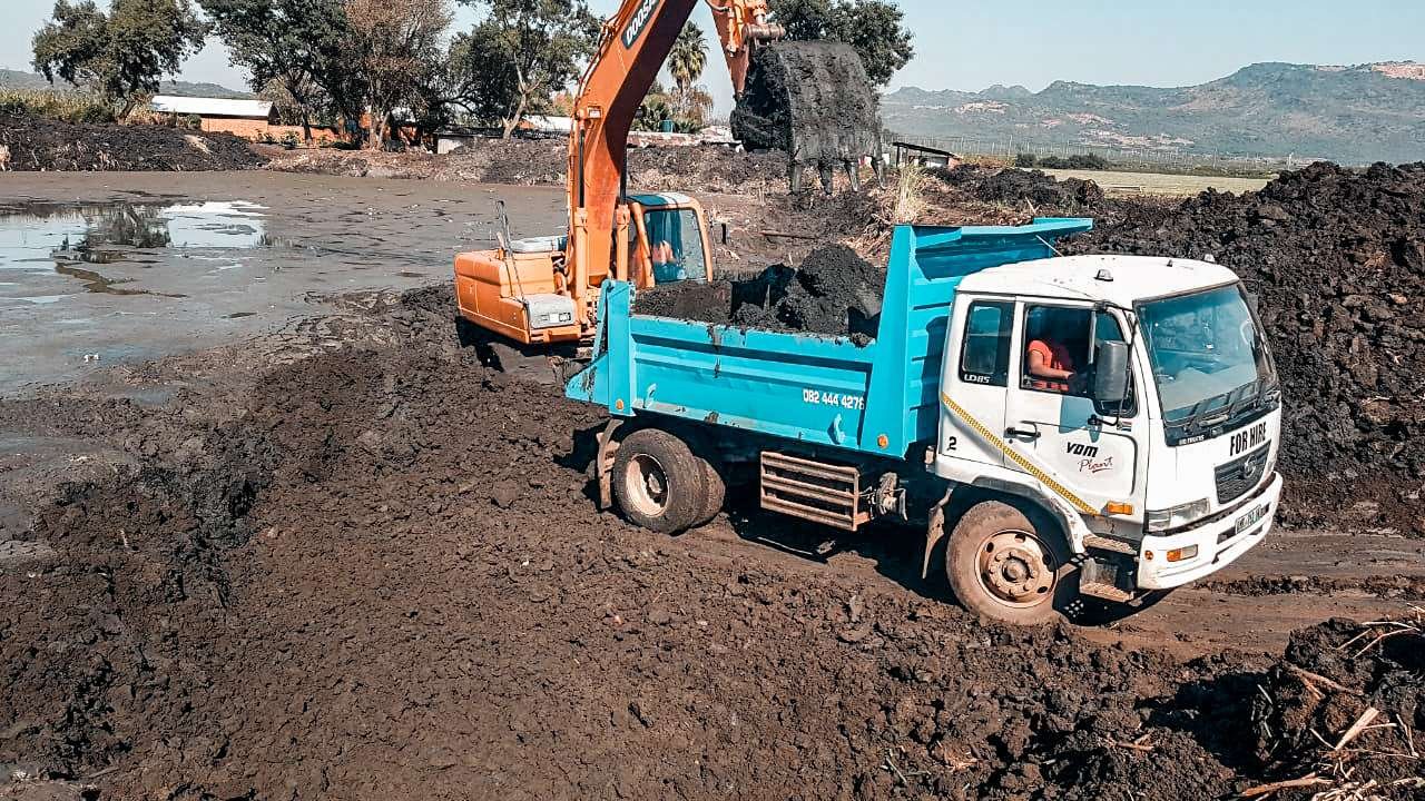 Excavator from a plant hire company loading soil onto a dump truck at a construction or excavation site with a scenic hilly backdrop.