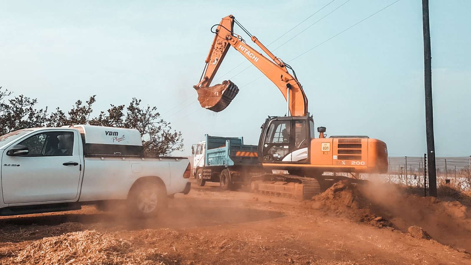 Heavy machinery at work: an excavator from VDM Plant Hire loading material into a dump truck at a construction or excavation site, with a pickup truck nearby. Dust is rising from the activity