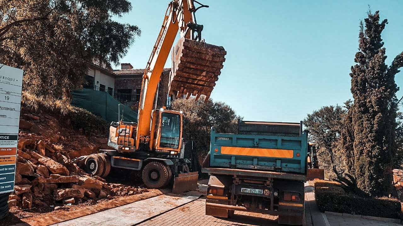 An excavator from VDM Plant Hire carefully loading large bricks or slabs onto a truck at a construction or renovation site, showcasing heavy machinery at work in an outdoor setting with greenery in the background